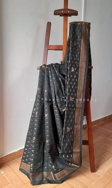 A black saree with metallic bronze zari motifs draped over a wooden display stand in an indoor setting with a light gray wall and wooden floor.
