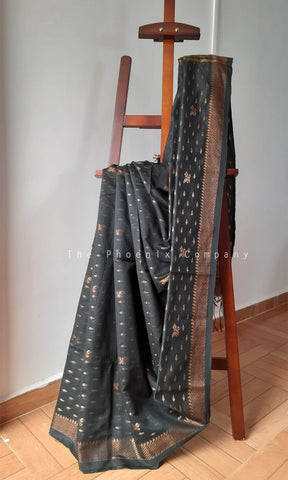 A black saree with metallic bronze zari motifs draped over a wooden display stand in an indoor setting with a light gray wall and wooden floor.
