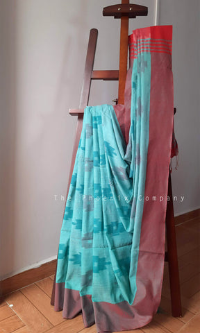 A draped sari in turquoise and dusty rose pink with a red border, displayed on a wooden easel in an indoor setting.