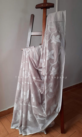 A sheer white saree with a light gray floral pattern draped over a wooden hanger in an indoor setting with a tiled floor and plain wall.
